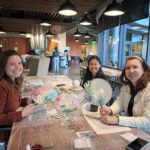 "Three women smiling while creating dry flower hand fans during a DIY craft workshop in a cozy, modern studio space."