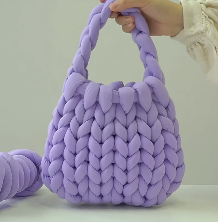 A handwoven velvet bag in soft lavender, featuring a chunky braided texture, held by a person in a light outfit.