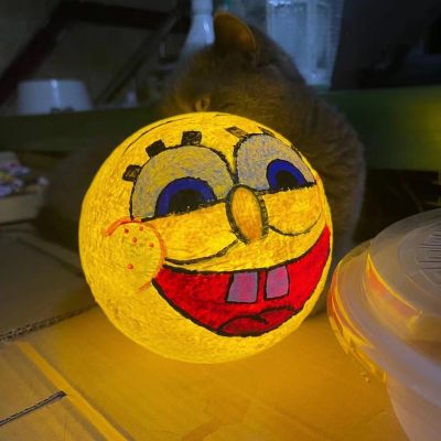 A glowing handmade moon light painted with a cheerful SpongeBob-style face, placed on a table in a dimly lit room.
