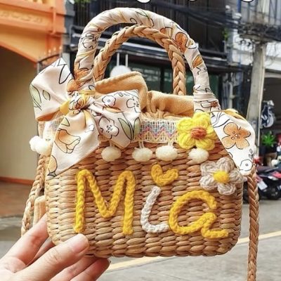 A personalized woven handbag decorated with yarn lettering, floral accents, pom-poms, and a patterned bow, held outdoors
