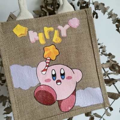 A hand-painted tote bag featuring a vibrant design of Kirby holding a star wand, with playful lettering and cloud accents.
