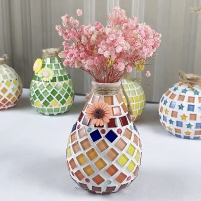 Handcrafted mosaic vase adorned with colorful tiles and pink dried flowers, surrounded by other vibrant mosaic vases on display.