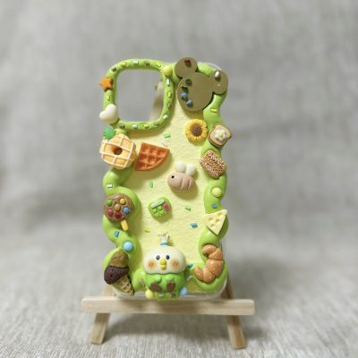 A handcrafted decoden phone case decorated with colorful, miniature 3D food and animal charms, arranged on a pastel yellow and green whipped cream-like background. The case is standing upright on a small wooden easel against a soft, neutral backdrop.